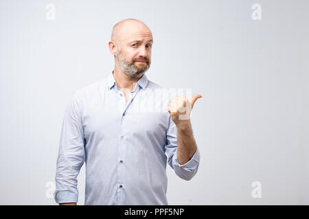 Gut aussehender bärtiger Mann in legere Kleidung zeigt awaywith Daumen auf Kamera schaut auf grauem Hintergrund Stockfoto