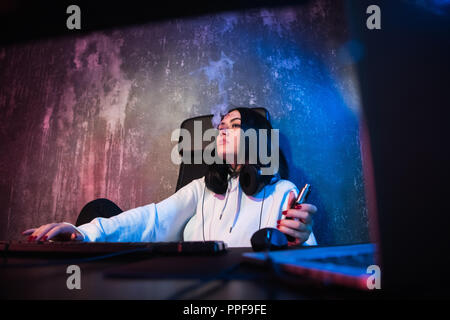 Junge hip brünette Frau mit Brille und Hoodie, Sitzen, Schreibtisch mit Computer, während Rauchen E-Zigarette Stockfoto