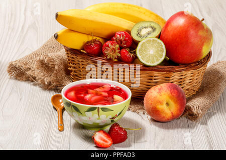 Cherry-Gelee mit Erdbeere Stücke in der Schale, frisch Erdbeeren, Nektarinen, Kalk, Kiwi, Banane, Apfel in einem Weidenkorb auf einem Holztisch Stockfoto