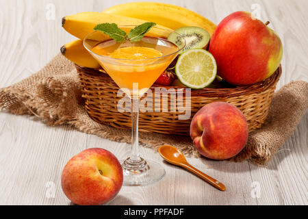 Orange Marmelade im Glas überstieg Minze, frische Erdbeeren, Nektarinen, Kalk, Kiwi, Banane, Apfel in einem Weidenkorb auf einem Holztisch Stockfoto