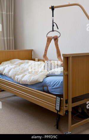 Alte Frau mit einer unheilbaren Krankheit, die versuchen, sich mit ihren extrem dünne und schwache Arme am Galgen der Krankenpflege Bett in ihrem Hospiz Zimmer zu ziehen Stockfoto