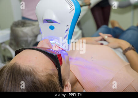 Close-up Portrait einer Patientin auf Zahnarzt in der Klinik. Zähne, die Verfahren mit UV-Licht UV-Lampe. Patienten mit Sicherheit Schutzbrille und Wange der Aufrollvorrichtung. Verfahren der professionellen Zähne, die Stockfoto