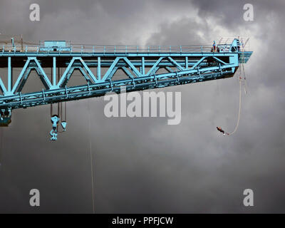 Gegen einen dunklen und grauer Himmel, ein Bungee Jumper hängt von der Ausleger des mächtigen Titan am Ufer des Flusses Clyde, Clydebank in der Nähe von Glasgow. Der Kran war einst Teil des berühmten John Brown Shipbuilding Yard und hat jetzt renoviert und ist ein beliebtes Reiseziel und Besucher, wo es einen atemberaubenden Blick von der Ausleger nach oben reisen der Lift dort zu erhalten. Stockfoto