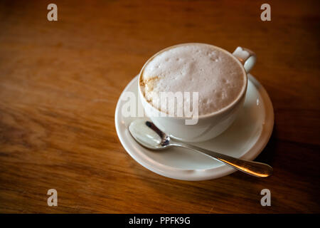 Frischen heißen Kaffee von oben Blick auf Holztisch. cappuchino Kaffee in weißen Ritual Zutaten Virtuelles Magazin Tasse und Untertasse mit silbernen Löffel. Minimale Zusammensetzung, hipster Vibes. Ansicht von oben, flach kopieren Platz für Ihren Text. Stockfoto