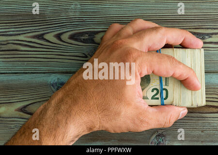 Mann hält eine gefaltete Wad von 20 Dollar Banknoten schützend in seiner Hand, er wird Sie in einem rustikalen Holztisch halten Stockfoto