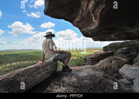 Ein männlicher Tourist, der unter einer Felskunstgalerie auf dem Injalak Hill sitzt und die herrliche Aussicht auf Arnhem Land, Northern Territory, Australien, betrachtet Stockfoto