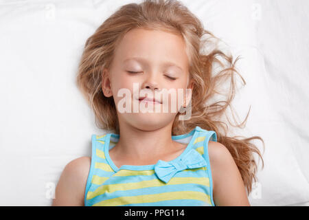 Gesunde ansprechende kleine Mädchen mit langen unordentlichen Haar, hält die Augen geschlossen, in pajamass gekleidet, hat angenehme dreans während der Nachtruhe, liegt am weißen Bett. Sm Stockfoto