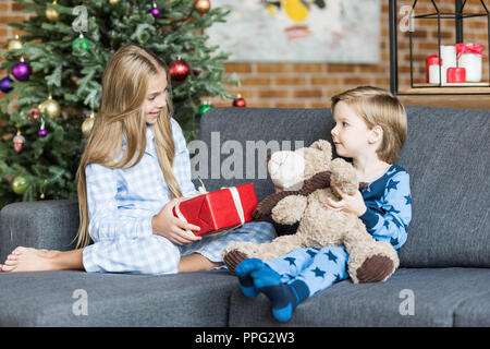 Niedliche happy Kids im Schlafanzug holding Teddybären und Weihnachtsgeschenk im Sofa sitzen und lächeln einander Stockfoto