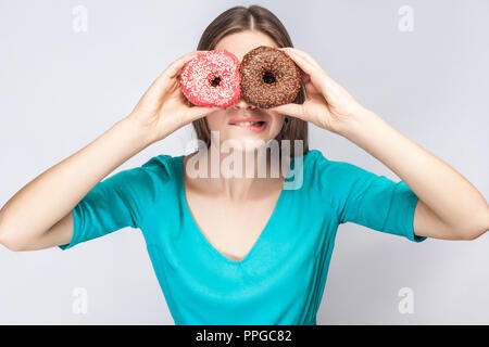 Junge Crazy Girl biss ein Lippen in blaue Bluse, die hielten, die Augen mit Rosa und Schokolade Donuts, schauen durch Donuts wie Glas Stockfoto