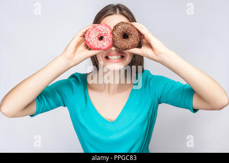 Positive junge lächelnde Mädchen in blau Bluse, die hielten, die Augen mit Rosa und Schokolade Donuts, schauen durch Donuts wie Gläser Stockfoto