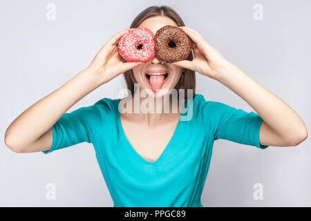 Positive Junge verrückte Mädchen in blau Bluse, die hielten, die Augen mit Rosa und Schokolade Donuts, schauen durch Donuts wie die Brille auf Stockfoto