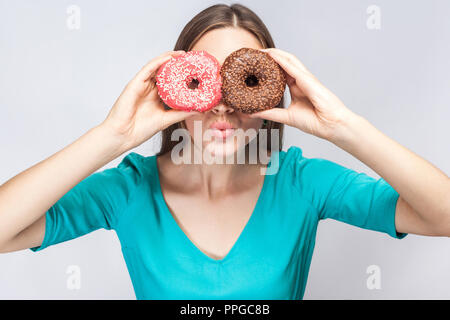 Positive junge schöne Mädchen in blau Bluse, die hielten, die Augen mit Rosa und Schokolade Donuts, schauen durch Donuts wie glasse Stockfoto