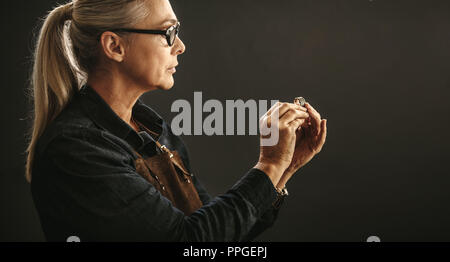 Ältere Frau Juwelier tragen Brillen und Schürze an der Qualität der Ring vor grauem Hintergrund. Goldschmied Prüfung der Ring für Mängel. Stockfoto