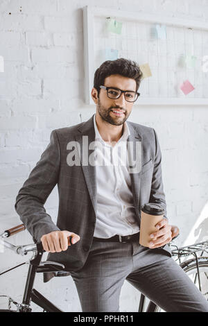 Lächelnd gut aussehender Geschäftsmann holding Tasse Kaffee und lehnte sich auf dem Fahrrad im Büro Stockfoto