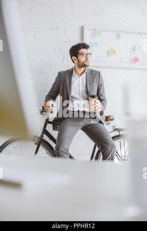 Gut aussehender Geschäftsmann sitzen auf dem Fahrrad und halten Kaffee im Büro gehen Stockfoto