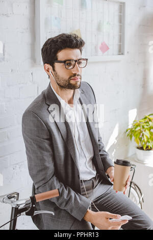 Geschäftsmann Musik hören mit Smartphone, Kaffee im Pappbecher und lehnte sich auf dem Fahrrad im Büro Stockfoto
