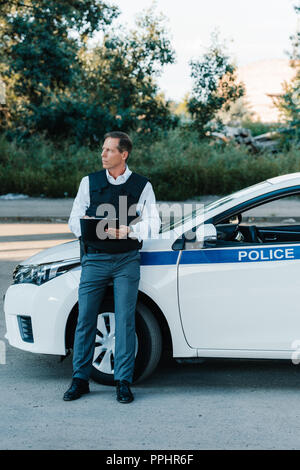 Reife männliche Polizisten in schusssicheren Weste schreiben in der Zwischenablage in der Nähe von Autos auf der Straße Stockfoto