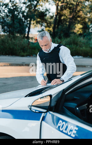 Reife männliche Polizisten schreiben in Zwischenablage auf Auto auf die Straße konzentriert Stockfoto