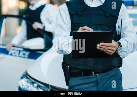 Zugeschnittenes Bild der männlichen Polizisten schreiben in Zwischenablage, während sein Kollege in der Nähe von Autos auf der Straße stehend Stockfoto