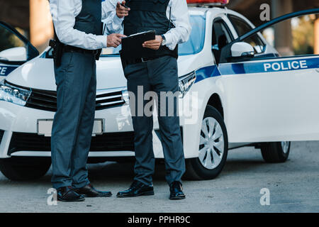 Teilweise mit Blick auf die männliche Polizisten mit Klemmbrett und zeigen mit dem Finger stehenden Kollegen in der Nähe von Autos auf der Straße zu Stockfoto