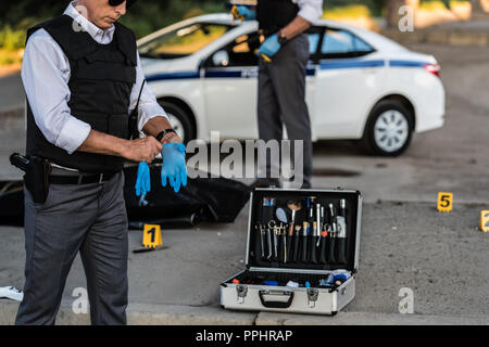 Teilweise mit Blick auf die männliche Polizisten bei Sonnenbrillen auf Latex Handschuhe, während seine Kollegen hinter am Tatort mit Leiche in body bag Stockfoto