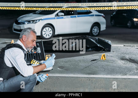 Seitenansicht des Polizisten Sammeln der Beweise in der Nähe von Cross Line am Tatort mit Leiche in body bag Stockfoto
