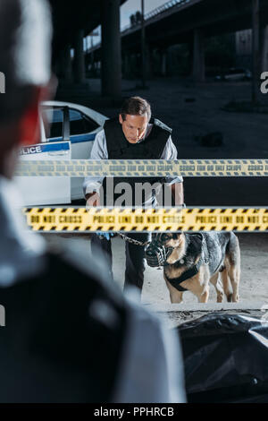 Teilweise mit Blick auf die männliche Polizisten stehen in der Nähe der Leiche in der Tasche, während seine Kollegen stehen mit Deutscher Schäferhund an der Leine am Tatort Stockfoto