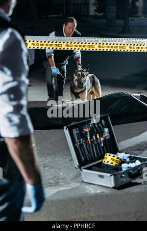 Teilweise mit Blick auf die Polizisten stehen in der Nähe von Case mit Untersuchung tools, während seine Kollegen stehen mit Deutscher Schäferhund an der Leine in der Nähe der Leiche bei c Stockfoto