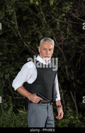 Schwere reifen Polizeioffizier in kugelsicheren Weste holding Gun in der Nähe von Forest Stockfoto