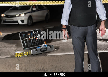 Zugeschnittenes Bild der Polizist stand in der Nähe von Cross Line und bei Untersuchung Werkzeuge in der Nähe der Leiche in Body Bag am Tatort Stockfoto