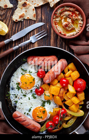 Blick von oben auf eine Pfanne mit Spiegeleier, gebratenen Würstchen, Ahorn verglaste Kürbis und Tomaten mit mediterraner dip Baba ganoush, pita Brea Stockfoto