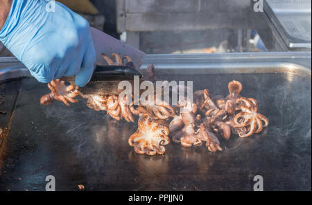 Küchenchef gegrillte Tintenfische. Kochvorführungen. Der Küchenchef flips kleine Kraken auf die Metallplatte des Grills. Dampf und Rauch aus dem Öl über Stockfoto