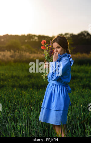 Junge Frau in blau und weiss gestreiftes Kleid hält einen Strauß Sommerblumen und Blick in die Kamera beim Stehen auf dem Gebiet der Grünen whea Stockfoto