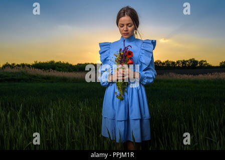 Junge Frau in blau und weiss gestreiftes Kleid, hält einen Strauß Sommerblumen mit geschlossenen Augen und ihr Kopf nach unten geneigt beim Stehen in Stockfoto