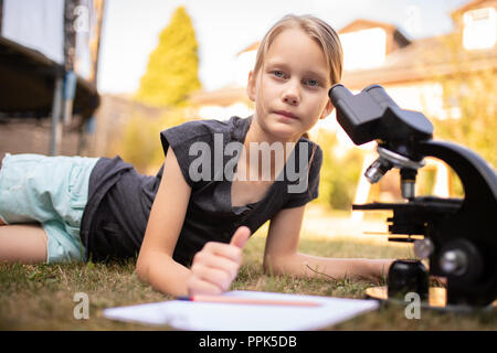 Ein 9 Jahre altes Mädchen ist, die auf dem Gras im Garten. Es hat ein Mikroskop und ein weißer Block vor. Das Mädchen sieht in die Kamera. Stockfoto