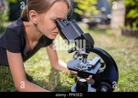 Ein 9 Jahre altes Mädchen liegt auf dem Rasen im Garten und schaut in das Okular eines Mikroskops. Stockfoto