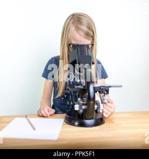 Ein 7-jähriges Mädchen schaut in ein Okular des Mikroskops und Notizen ihre Beobachtungen auf ein Blatt Papier. Vor weißem Hintergrund Stockfoto