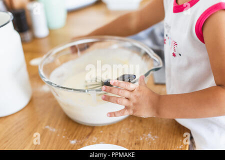 Kleines Mädchen Zubereitung Teig für Pfannkuchen in der Küche. Konzept der Lebensmittelzubereitung, weiße Küche für den Hintergrund. Casual Lifestyle Foto serie in re Stockfoto