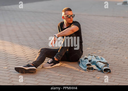 Stilvolle gutaussehenden jungen Mann mit Brille in schwarzer Kleidung mit Sneakers sitzt auf der Straße Stockfoto
