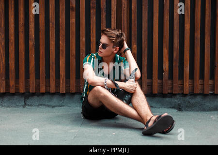 Stilvolle junger stattlicher Mann mit Sonnenbrille in trendigen Sommer Kleidung mit Schuhen in der Nähe von eine hölzerne Wand saß Stockfoto