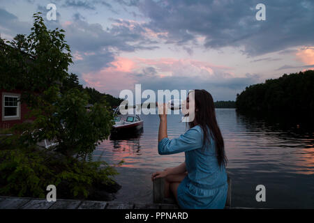 Junge brünette Frau auf einem lakeside Dock bei Sonnenuntergang sitzen bei einem Glas Rotwein. Stockfoto