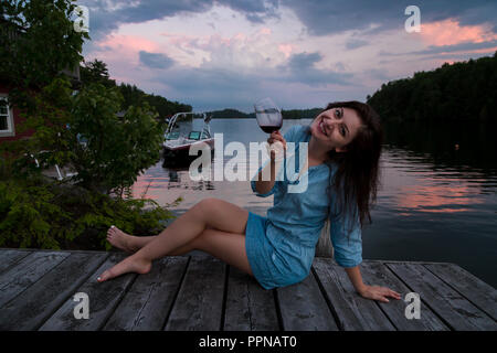 Junge brünette Frau auf einem lakeside Dock bei Sonnenuntergang sitzen bei einem Glas Rotwein. Stockfoto