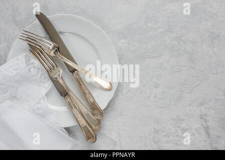 Silber Besteck auf einem Porzellanteller und weiß Serviette mit belgischen Spitze sind auf dem Hintergrund von grauem Beton Oberfläche, mit copy-Raum Stockfoto