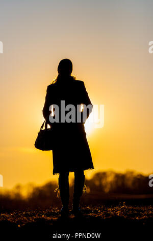 Die Silhouette eines jungen Mädchens in einen Mantel, Stiefel und eine Tasche im Frühjahr Wald bei Sonnenuntergang im April. Hintergrundbeleuchtung Sonnenlicht. Vor dem Hintergrund einer Wolkenlosen Stockfoto