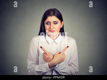 Junge unsichere Frau suchen betonte gleichzeitig, mit den Fingern in entgegengesetzte Richtungen auf grauem Hintergrund Stockfoto