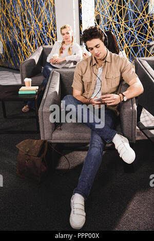 Junge Unternehmer in Kopfhörer mit Smartphone sitzt auf einem Sessel und Geschäftsfrau hinter mit Kaffee an modernen coworking Büro Stockfoto