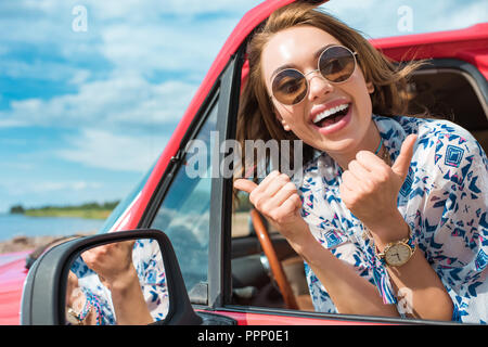 Neugierige junge Frau in Sonnenbrille im Auto sitzen und Daumen hoch während der Reise Stockfoto