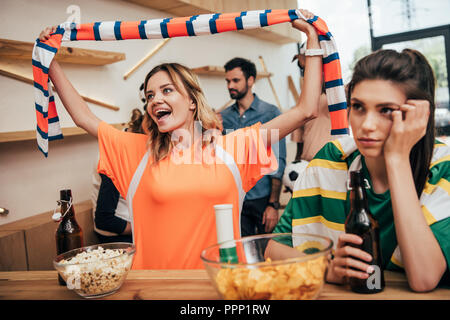 Neugierige junge Frau in orange Fan t-shirt Feiern und hält das Gebläse Schal über dem Kopf, während Ihr umgekippt Freundin in Grün t-shirt Sitzen mit Bier Stockfoto