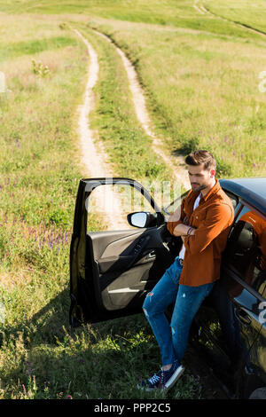 Erhöhten Blick auf jungen Mann mit gekreuzten Händen neben seinem Auto steht im Feld Stockfoto