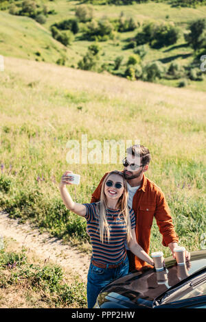 Hohe Betrachtungswinkel von stilvollen Paar in Sonnenbrille mit Kaffeetassen, selfie auf dem Smartphone in der Nähe von Auto auf ländlichen Wiese Stockfoto